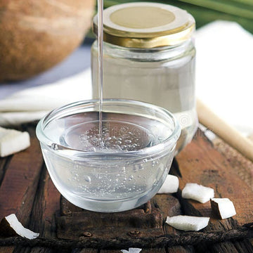 Coconut oil being poured from a jar into a glass bowl on a wooden surface with coconuts and a spoon.